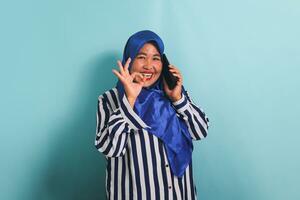 un emocionado de edad mediana asiático mujer en un azul hijab y un a rayas camisa es haciendo un Okay gesto o aprobación firmar mientras teniendo un teléfono conversación. ella es aislado en un azul antecedentes foto