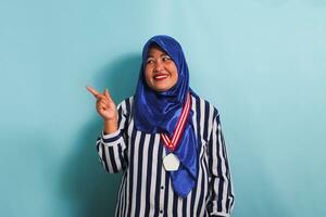 A happy middle-aged Asian woman, wearing a blue hijab and a striped shirt, is pointing her finger to the left towards the copyspace. She is wearing a medal and is isolated on a blue background photo