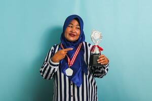 un contento de edad mediana asiático mujer de negocios en un azul hiyab, a rayas camisa, y medalla es señalando un plata trofeo, celebrando su éxito y logro, aislado en un azul antecedentes foto