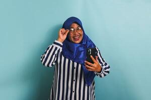 An excited middle-aged Asian woman in a blue hijab, wearing eyeglasses and a striped shirt, is laughing while looking at the camera and holding a mobile phone. She is isolated on a blue background. photo