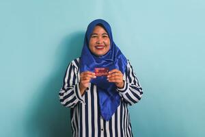 Excited Middle-aged Asian woman in blue hijab and striped shirt showing, presenting credit card for paying online business, isolated over blue background. photo