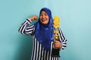 un orgulloso de edad mediana asiático mujer de negocios en un azul hijab y un a rayas camisa es haciendo un fuerte gesto mientras participación un oro trofeo, celebrando su éxito. ella es aislado en un azul antecedentes. foto