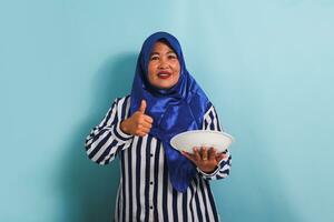 A middle-aged Asian woman in a blue hijab and a striped shirt is giving a thumbs up while holding an empty white plate. She is isolated on a blue background photo