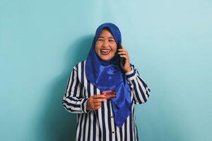 An excited middle-aged Asian woman in a blue hijab and a striped shirt is talking on the phone and showing a credit card. She is isolated on a blue background photo