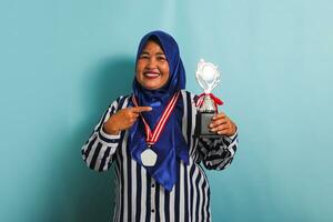 A happy middle-aged Asian businesswoman in a blue hijab, striped shirt, and medal is pointing a silver trophy, celebrating her success and achievement, isolated on a blue background photo