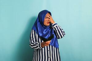A shocked middle-aged Asian woman in a blue hijab and a striped shirt is peeking through her fingers into her empty wallet with a shocked and surprised expression. She is isolated on a blue background photo