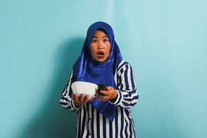 A shocked middle-aged Asian woman in a blue hijab is watching television, pointing the TV remote controller towards the camera, and switching channels with an open mouth, Isolated on a blue background photo
