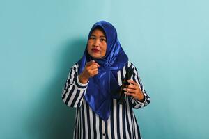 A middle-aged Asian woman in a blue hijab and a striped shirt is pointing at the camera while holding an empty wallet and looking frustrated. She is isolated on a blue background photo