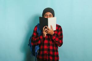 A young Asian man with a backpack, wearing a beanie hat and a red plaid flannel shirt, is covering his face with a book while standing against a blue background photo