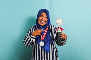 un contento de edad mediana asiático mujer de negocios en un azul hiyab, a rayas camisa, y medalla es señalando un plata trofeo, celebrando su éxito y logro, aislado en un azul antecedentes foto