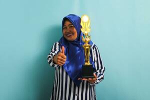 un orgulloso de edad mediana asiático mujer de negocios en un azul hijab y un a rayas camisa es haciendo un pulgar arriba gesto mientras participación un oro trofeo, celebrando su éxito. ella es aislado en un azul antecedentes. foto