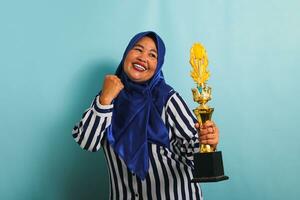 un orgulloso de edad mediana asiático mujer de negocios en un azul hijab y un a rayas camisa es haciendo un ganador gesto mientras participación un oro trofeo, celebrando su éxito. ella es aislado en un azul antecedentes. foto
