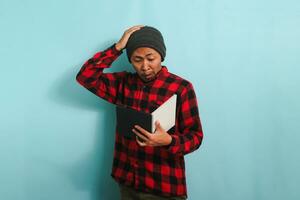 Confused young Asian male student with beanie hat and red plaid flannel shirt looks shocked scratching his head, has forgotten to learn and is not prepared for exams, isolated on blue background photo