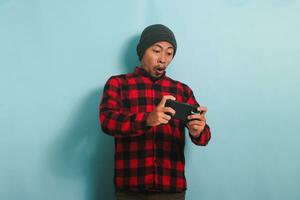 emocionado joven asiático hombre con un gorro sombrero y un rojo tartán franela camisa es jugando un en línea juego en su teléfono inteligente, aislado en un azul antecedentes foto