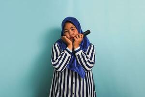 A bored middle-aged Asian woman in a blue hijab and a striped shirt is watching a boring TV show and switching channels with a remote control. She is isolated on a blue background. photo