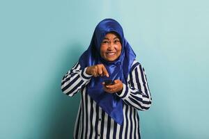 An annoyed middle-aged Asian woman in a blue hijab and a striped shirt is trying to switch channels while watching a movie on TV. She is isolated on a blue background photo