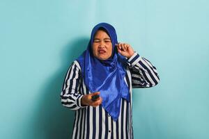 An annoyed middle-aged Asian woman in a blue hijab and a striped shirt is trying to switch channels while watching a movie on TV. She is isolated on a blue background photo