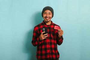 Happy young Asian man with a beanie hat and a red plaid flannel shirt is holding a smartphone and a banking credit card, doing online shopping, looking at camera, isolated on a blue background. photo