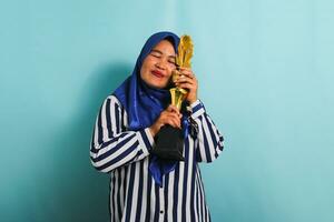 un emocionado de edad mediana asiático mujer de negocios en un azul hijab y un a rayas camisa es abrazando un oro trofeo, celebrando su éxito y logro. ella es aislado en un azul antecedentes foto