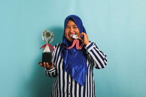 un contento de edad mediana asiático mujer de negocios en un azul hijab y un a rayas camisa es mordiendo un medalla mientras participación un plata trofeo, celebrando su éxito y logro, aislado en un azul antecedentes foto