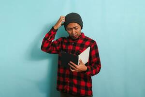 Confused young Asian male student with beanie hat and red plaid flannel shirt looks shocked scratching his head, has forgotten to learn and is not prepared for exams, isolated on blue background photo