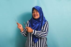 An excited middle-aged Asian woman in a blue hijab and a striped shirt is pointing to the left copyspace with a spoon and fork. She is isolated on a blue background photo