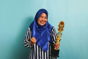 un orgulloso de edad mediana asiático mujer de negocios en un azul hijab y un a rayas camisa es haciendo un ganador gesto mientras participación un oro trofeo, celebrando su éxito. ella es aislado en un azul antecedentes. foto