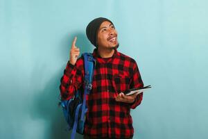 An Excited smiling young Asian student with a beanie hat and a red plaid flannel shirt, wearing a backpack, is pointing finger up at copy space while standing against a blue background photo