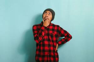 Thoughtful Young Asian man holds his chin, making plans, isolated on a blue background photo