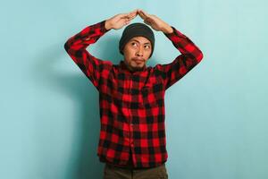 Pensive young millennial Asian man making a roof gesture above his head, isolated on blue background photo