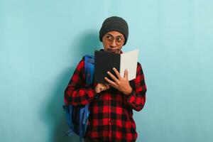 Pensive young Asian student man is thinking while holding book and standing against blue background photo