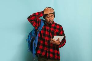 Surprised young Asian student appears unhappy with his hand on his head, isolated on blue background photo