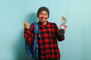 Excited young Asian man student makes YES gesture while holding trophy, isolated on blue background photo