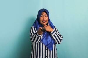A shocked middle-aged Asian woman in a blue hijab is watching television, pointing the TV remote controller towards the camera, and switching channels with an open mouth, Isolated on a blue background photo