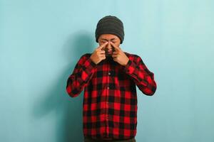 Young Asian man rubbing his swollen eyes due to dust allergy and itching isolated on blue background photo