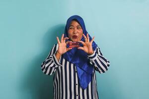 Excited Middle-aged Asian woman in blue hijab and striped shirt showing, presenting credit card for paying online business, isolated over blue background. photo