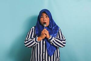 A hungry middle-aged Asian woman in a blue hijab and a striped shirt has a spoon in her mouth as she looks at Camera. She is thinking about what to eat. She is isolated on a blue background photo