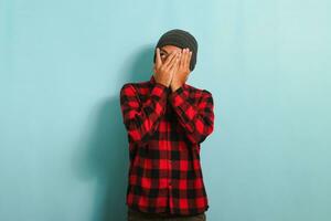 Embarrassed young Asian man facepalms, covering his face with his hand, isolated on blue background photo