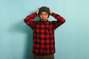 Unhappy young Asian man grabs his head feeling anxious while standing against a blue background photo