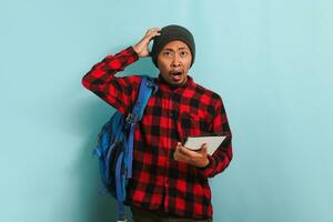 A surprised young Asian student with a beanie hat and a red plaid flannel shirt, wearing a backpack, scratching his head appears unhappy and confused, isolated on a blue background photo