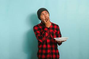 Pensive Young Asian man thinking about what to eat while holding plate, isolated on blue background photo