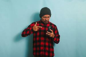 Angry young Asian man with a beanie hat and a red plaid flannel shirt is yelling and pointing at his phone, isolated on a blue background photo