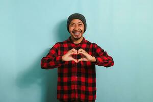 Smiling young Asian man shows a love gesture with his hand while standing against a blue background photo