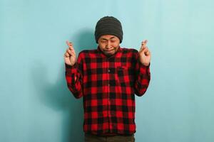 Hopeful young Asian man makes a finger crossed gesture, closes his eyes, isolated on blue background photo