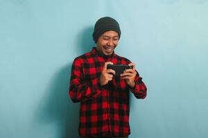 emocionado joven asiático hombre con un gorro sombrero y un rojo tartán franela camisa es jugando un en línea juego en su teléfono inteligente, aislado en un azul antecedentes foto