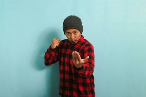 Angry young Asian man is ready to fight, with a defensive fist gesture, isolated on blue background photo