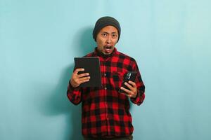 Confused young Asian male student with beanie hat and red plaid flannel shirt looks shocked, has forgotten to learn and is not prepared for exams, isolated on blue background photo