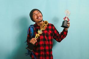 contento joven asiático hombre estudiante levantamiento arriba su trofeos, aislado en azul antecedentes. foto