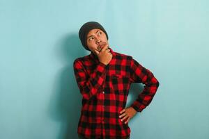 Pensive young Asian man keeps his hand on his chin, looking at copy space, isolated on blue backdrop photo