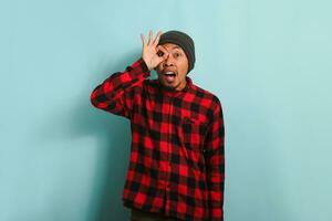 Excited young Asian man makes the OK sign gesture like binoculars, isolated on blue background photo